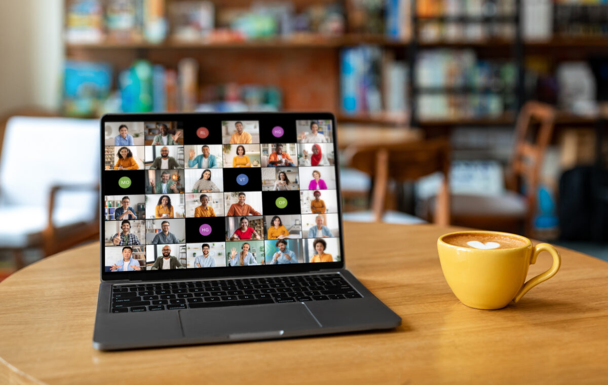A laptop computer sits on a wooden table in a cafe. The laptop screen displays a video conference meeting with multiple participants from different backgrounds.