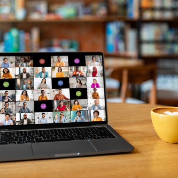 A laptop computer sits on a wooden table in a cafe. The laptop screen displays a video conference meeting with multiple participants from different backgrounds.