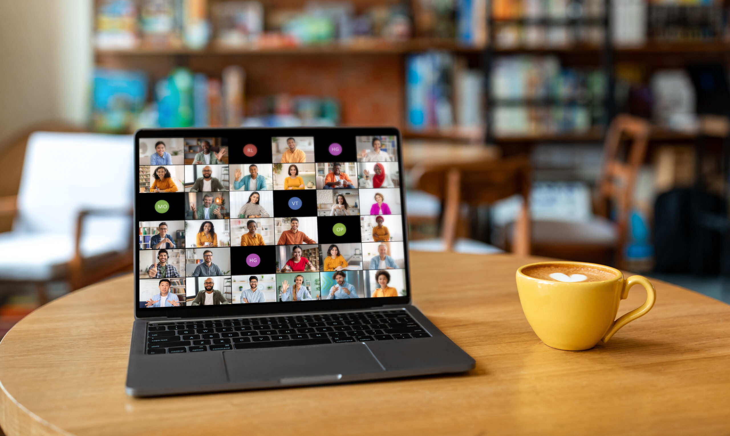 A laptop computer sits on a wooden table in a cafe. The laptop screen displays a video conference meeting with multiple participants from different backgrounds.